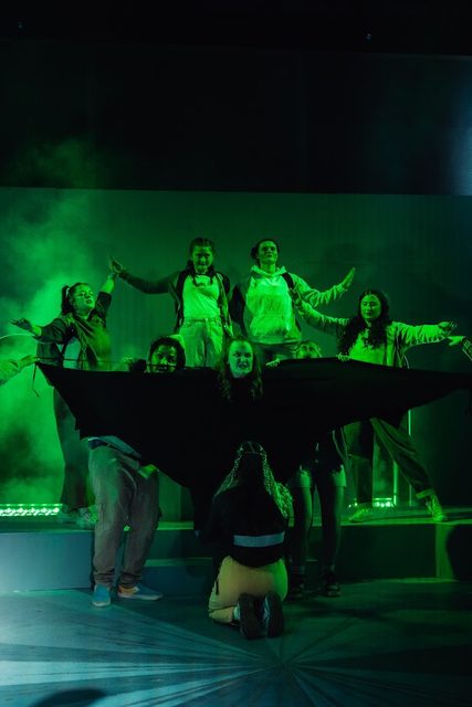 Ensemble scene lit in green, cast gathered around an upturned boat